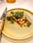 A beautifully arranged gourmet dish sits on a textured ceramic plate. The dish features a colorful assortment of vegetables, vibrant greens, and delicate edible flowers. A polished knife with a decorative handle is placed next to the plate, suggesting an emphasis on presentation and fine dining.