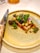 A beautifully arranged gourmet dish sits on a textured ceramic plate. The dish features a colorful assortment of vegetables, vibrant greens, and delicate edible flowers. A polished knife with a decorative handle is placed next to the plate, suggesting an emphasis on presentation and fine dining.