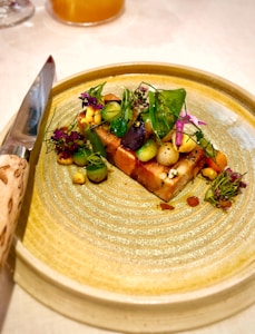 A beautifully arranged gourmet dish sits on a textured ceramic plate. The dish features a colorful assortment of vegetables, vibrant greens, and delicate edible flowers. A polished knife with a decorative handle is placed next to the plate, suggesting an emphasis on presentation and fine dining.