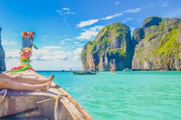 A traditional wooden boat is gently floating on clear turquoise waters, surrounded by towering limestone cliffs covered in lush green vegetation. The sky is bright blue with scattered white clouds, and another boat can be seen in the distance. On the boat's prow, there are colorful decorations and a person's legs resting comfortably, suggesting a relaxed atmosphere.