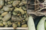 A wooden crate filled with rough, misshapen potatoes, surrounded by other wooden crates containing squash and eggplants. The potatoes have visible dirt patches, and dried straw is scattered around the edges, suggesting a rustic or farm setting.