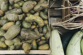 A wooden crate filled with rough, misshapen potatoes, surrounded by other wooden crates containing squash and eggplants. The potatoes have visible dirt patches, and dried straw is scattered around the edges, suggesting a rustic or farm setting.