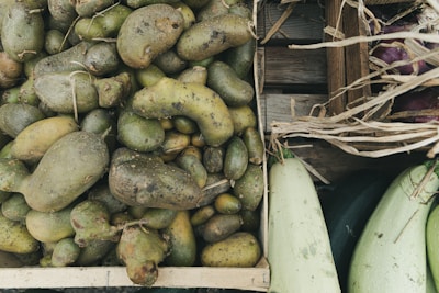A wooden crate filled with rough, misshapen potatoes, surrounded by other wooden crates containing squash and eggplants. The potatoes have visible dirt patches, and dried straw is scattered around the edges, suggesting a rustic or farm setting.