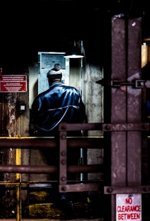 A person wearing a jacket and a hat is standing in a dimly lit industrial environment. The walls are aged and there is a sign about fire suppression systems. A lamp hangs overhead illuminating a map or diagram on the wall.