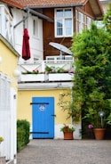 A charming residential setting features two-story buildings with wooden and white walls. The ground level showcases a bright yellow wall adorned with a striking blue door, marked by a bicycle symbol. Above, a balcony with white railings is decorated with colorful flower boxes. A red umbrella and a satellite dish add detail to the upper level. Verdant shrubs and potted plants contribute to the natural ambiance.