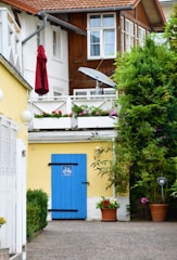 A charming residential setting features two-story buildings with wooden and white walls. The ground level showcases a bright yellow wall adorned with a striking blue door, marked by a bicycle symbol. Above, a balcony with white railings is decorated with colorful flower boxes. A red umbrella and a satellite dish add detail to the upper level. Verdant shrubs and potted plants contribute to the natural ambiance.