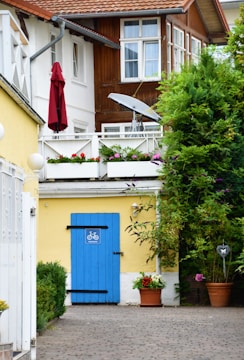 A charming residential setting features two-story buildings with wooden and white walls. The ground level showcases a bright yellow wall adorned with a striking blue door, marked by a bicycle symbol. Above, a balcony with white railings is decorated with colorful flower boxes. A red umbrella and a satellite dish add detail to the upper level. Verdant shrubs and potted plants contribute to the natural ambiance.