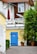 A charming residential setting features two-story buildings with wooden and white walls. The ground level showcases a bright yellow wall adorned with a striking blue door, marked by a bicycle symbol. Above, a balcony with white railings is decorated with colorful flower boxes. A red umbrella and a satellite dish add detail to the upper level. Verdant shrubs and potted plants contribute to the natural ambiance.