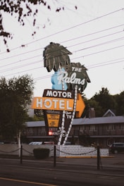A retro-style motel sign reads 'The Palms Motor Hotel' with neon lights and features palm tree illustrations. The sign offers amenities like free TV and continental rates. The setting includes power lines above and a glimpse of the motel building in the background, creating a nostalgic roadside atmosphere.