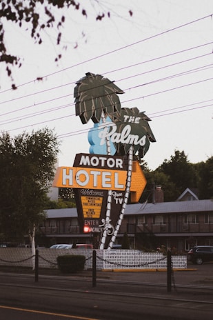 A retro-style motel sign reads 'The Palms Motor Hotel' with neon lights and features palm tree illustrations. The sign offers amenities like free TV and continental rates. The setting includes power lines above and a glimpse of the motel building in the background, creating a nostalgic roadside atmosphere.