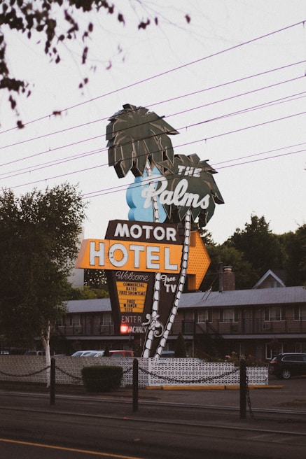 A retro-style motel sign reads 'The Palms Motor Hotel' with neon lights and features palm tree illustrations. The sign offers amenities like free TV and continental rates. The setting includes power lines above and a glimpse of the motel building in the background, creating a nostalgic roadside atmosphere.