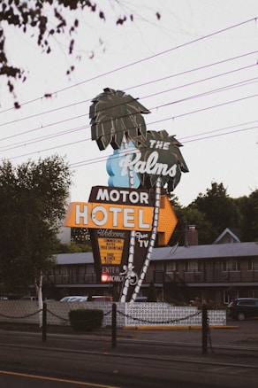 A retro-style motel sign reads 'The Palms Motor Hotel' with neon lights and features palm tree illustrations. The sign offers amenities like free TV and continental rates. The setting includes power lines above and a glimpse of the motel building in the background, creating a nostalgic roadside atmosphere.