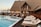 A luxurious wooden resort with a thatched roof, featuring a pool surrounded by lounge chairs and a wooden deck. There are a few people relaxing and walking around the area, and the setting sun casts a warm glow on the building and surroundings.