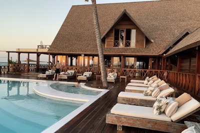 A luxurious wooden resort with a thatched roof, featuring a pool surrounded by lounge chairs and a wooden deck. There are a few people relaxing and walking around the area, and the setting sun casts a warm glow on the building and surroundings.