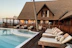 A luxurious wooden resort with a thatched roof, featuring a pool surrounded by lounge chairs and a wooden deck. There are a few people relaxing and walking around the area, and the setting sun casts a warm glow on the building and surroundings.