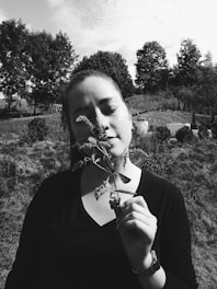 A woman stands in a grassy outdoor setting, eyes closed, holding a sprig of flowers close to her face. Trees and a partially cloudy sky can be seen in the background, creating a serene and natural atmosphere.