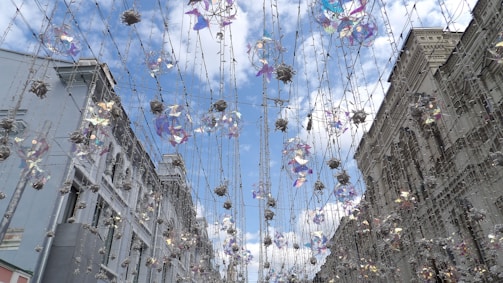 Strings of decorative lights and hanging ornaments stretch across a city street between two ornate buildings. The decorations feature iridescent and floral designs, creating a festive atmosphere under a bright, partially cloudy sky.