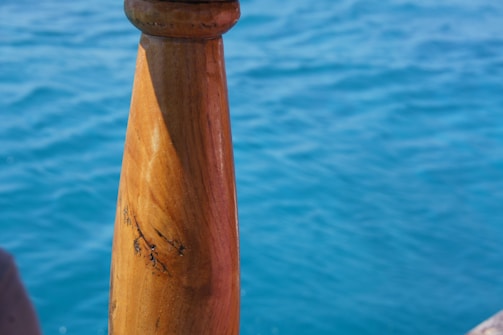 A close-up view of a wooden handrail or spindle with a polished finish, positioned against a backdrop of clear blue water. The wood grain and texture are visible, with sunlight casting a natural glow.
