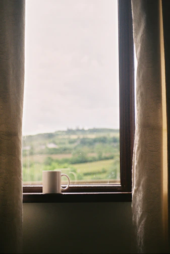 A vibrant white mug with the BuildIt Prints logo, filled with steaming coffee on a sunny windowsill.