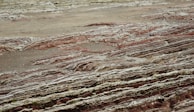 Layers of sedimentary rock formations with varying colors and textures are exposed on the surface. The patterns consist of red, brown, and gray hues, reminiscent of geological activity and erosion over time. The ground has a rugged, uneven appearance with visible stratification, indicating a rich geological history.