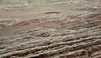 Layers of sedimentary rock formations with varying colors and textures are exposed on the surface. The patterns consist of red, brown, and gray hues, reminiscent of geological activity and erosion over time. The ground has a rugged, uneven appearance with visible stratification, indicating a rich geological history.