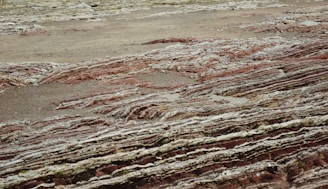 Layers of sedimentary rock formations with varying colors and textures are exposed on the surface. The patterns consist of red, brown, and gray hues, reminiscent of geological activity and erosion over time. The ground has a rugged, uneven appearance with visible stratification, indicating a rich geological history.