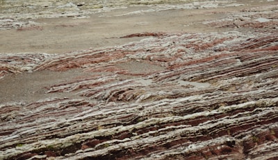 Layers of sedimentary rock formations with varying colors and textures are exposed on the surface. The patterns consist of red, brown, and gray hues, reminiscent of geological activity and erosion over time. The ground has a rugged, uneven appearance with visible stratification, indicating a rich geological history.
