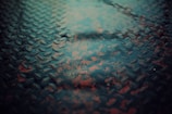 A textured metal surface with a diamond plate pattern is partially covered by raindrops, creating reflective patches accented by weathered red paint peeking through the dark metal.