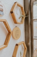 Wooden hexagonal shelves are mounted on a white textured wall. Inside the shelves are small plants and a piece of paper. In the center, there is an embroidery hoop with a simple line drawing of a face. The shelves and decorations are arranged next to a large window, allowing natural light to softly illuminate the scene.