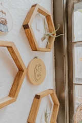 Wooden hexagonal shelves are mounted on a white textured wall. Inside the shelves are small plants and a piece of paper. In the center, there is an embroidery hoop with a simple line drawing of a face. The shelves and decorations are arranged next to a large window, allowing natural light to softly illuminate the scene.