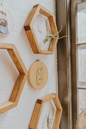 Wooden hexagonal shelves are mounted on a white textured wall. Inside the shelves are small plants and a piece of paper. In the center, there is an embroidery hoop with a simple line drawing of a face. The shelves and decorations are arranged next to a large window, allowing natural light to softly illuminate the scene.