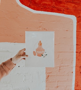 A hand holds up an artwork in front of a textured wall. The artwork depicts a vintage perfume bottle labeled with the word 'positivity' and small floral details. The wall has a peach and white abstract mural design.