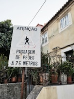 A street sign alerts pedestrians to a crossing 75 meters ahead. The sign, written in Portuguese, is placed in front of a building with potted plants and a wooden fence. The weathered building features a cross and has a classic architectural style.