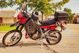 A motorcycle with a protective windshield and a storage box is parked in a sunny, urban environment. It features a red and black color scheme, with knobby tires indicating it is suitable for off-road travel. The background shows a small commercial area with buildings and a tree providing some shade.