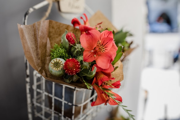A bouquet of vibrant flowers with red blooms and green foliage is wrapped in brown paper. The arrangement is placed inside a wire basket against a blurred background.