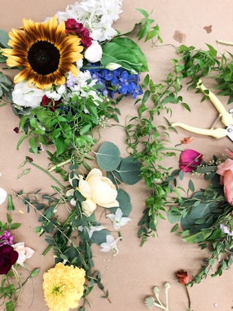 A collection of various flowers and green foliage is spread out on a brown surface. The arrangement includes a prominent sunflower with bright yellow petals, a mix of other flowers such as red, pink, and white roses, blue delphiniums, and yellow marigolds. Scattered among the flowers are different types of green leaves and vines. A pair of yellow garden shears is positioned to the right.