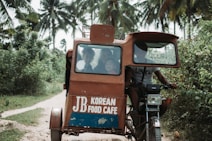 A motorized tricycle is navigating a narrow dirt path surrounded by lush greenery and palm trees. The vehicle has a small cabin with passengers visible through the front window and signage on the body displaying 'JB Korean Food Cafe.' The tricycle is being driven by a person who is partially visible.
