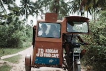 A motorized tricycle is navigating a narrow dirt path surrounded by lush greenery and palm trees. The vehicle has a small cabin with passengers visible through the front window and signage on the body displaying 'JB Korean Food Cafe.' The tricycle is being driven by a person who is partially visible.