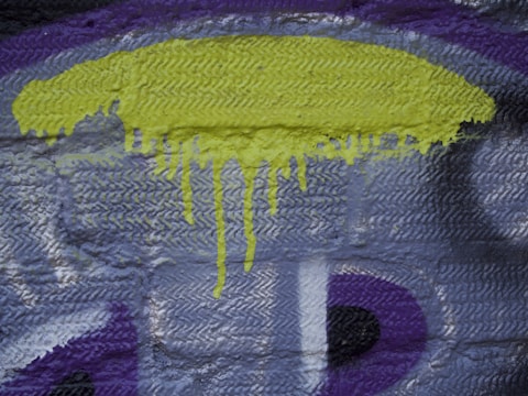 Graffiti on a textured brick wall, featuring a prominent yellow drip-like paint at the top center. The background consists of various shades of gray and purple, with visible brush strokes and patterns.