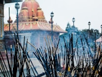 A collection of incense sticks, some still burning, stand in the foreground with visible smoke rising from them. Beyond the incense is a temple with a large, ornately decorated dome featuring gold embellishments atop a rich red structure. Several lampposts are evenly spaced along the pathway around the temple, with trees and shrubbery in the background.
