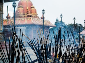 A collection of incense sticks, some still burning, stand in the foreground with visible smoke rising from them. Beyond the incense is a temple with a large, ornately decorated dome featuring gold embellishments atop a rich red structure. Several lampposts are evenly spaced along the pathway around the temple, with trees and shrubbery in the background.