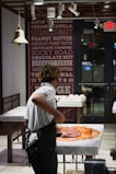 A person wearing a white shirt and apron is working with a large amount of chocolate on a marble counter in a well-lit shop or kitchen. The background features a wall decorated with text advertising various flavors and an 'Exit' sign above a door.