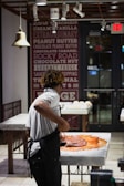 A person wearing a white shirt and apron is working with a large amount of chocolate on a marble counter in a well-lit shop or kitchen. The background features a wall decorated with text advertising various flavors and an 'Exit' sign above a door.
