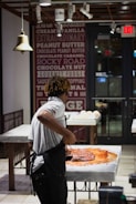 A person wearing a white shirt and apron is working with a large amount of chocolate on a marble counter in a well-lit shop or kitchen. The background features a wall decorated with text advertising various flavors and an 'Exit' sign above a door.