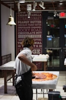 A person wearing a white shirt and apron is working with a large amount of chocolate on a marble counter in a well-lit shop or kitchen. The background features a wall decorated with text advertising various flavors and an 'Exit' sign above a door.