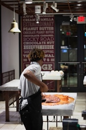 A person wearing a white shirt and apron is working with a large amount of chocolate on a marble counter in a well-lit shop or kitchen. The background features a wall decorated with text advertising various flavors and an 'Exit' sign above a door.