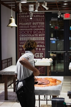 A person wearing a white shirt and apron is working with a large amount of chocolate on a marble counter in a well-lit shop or kitchen. The background features a wall decorated with text advertising various flavors and an 'Exit' sign above a door.