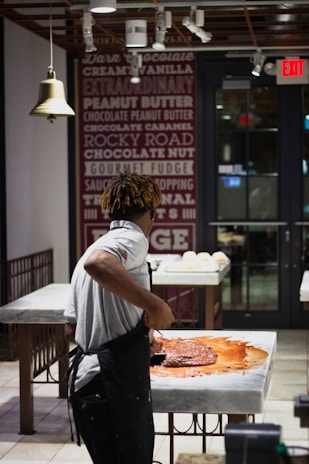 A person wearing a white shirt and apron is working with a large amount of chocolate on a marble counter in a well-lit shop or kitchen. The background features a wall decorated with text advertising various flavors and an 'Exit' sign above a door.