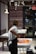 A person wearing a white shirt and apron is working with a large amount of chocolate on a marble counter in a well-lit shop or kitchen. The background features a wall decorated with text advertising various flavors and an 'Exit' sign above a door.