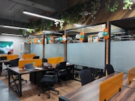 An open office space with wooden flooring and green plants hanging from the ceiling. There are multiple workstations with black chairs and orange partitions. Balloons in orange, white, and green are used for decoration, suggesting a festive or themed setup. Laptops and office supplies can be seen on the desks, and there is a painting on the wall to the left.